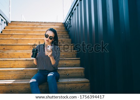 Millennial blogger in stylish sunglasses writing email message for send during mobility browsing, Caucasian hipster girl with coffee to go resting at urban setting using cellular for creating content