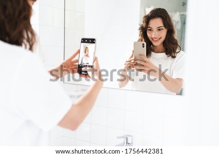 Photo of cheerful beautiful woman smiling and taking selfie on cellphone while looking at mirror at bathroom