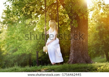 A girl in a white dress rides on a swing in nature and the horse grazes in the meadow. Pastoral paradise picture.