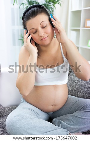 Pregnant woman sitting on white sofa at home and listening music in headphones 