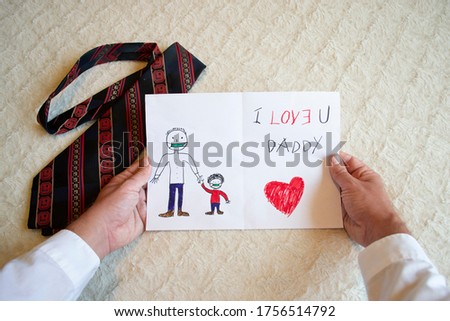 Man hands holding greeting card drawn on paper by his child for father's day representing father holding hand with son daughter and writing I Love U you Daddy with a red black striped tie holiday
