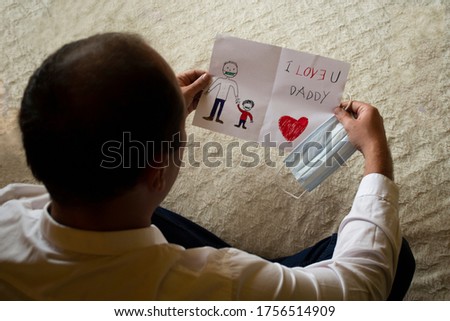 Man blurred holding sharped mask and greeting card drawn on paper by his child for father's day representing father holding hand with son daughter and writing I Love U you Daddy celebrating holiday