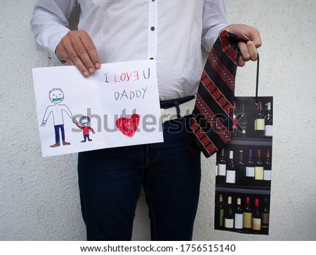 Man holding greeting card with writing happy fathers day daddy and son wearing face mask, red heart, red dark striped tie and bottle gift bag, happy with the presents he got from his children
