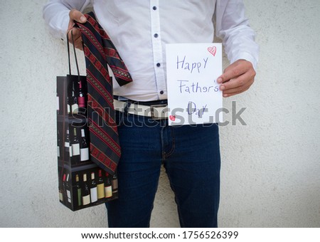 Man holding greeting card with writing happy fathers day, red dark striped tie and bottle gift bag, happy with the presents he got from his children, standing before a white background