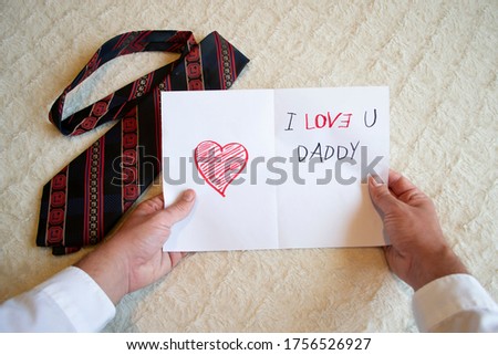 Man holding in the hands a greeting card drawn by his child for father's day during coronavirus pandemic representing red heart with writing I love U you Daddy in the background with a red dark tie