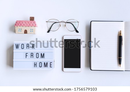 Flat lay of white desk with notebook, glasses, smartphone, home model and work from home word on lightbox. Copy space, top view. Quarantine, new normal, home office  concept.