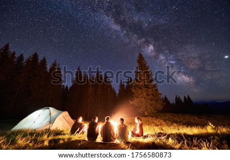 Night summer camping in the mountains, spruce forest on background, sky with stars and milky way. Back view group of five tourists having a rest together around campfire, enjoying fresh air near tent.