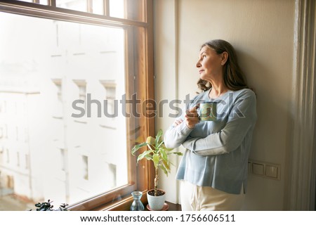 Portrait of thoughtful retired mature woman in casual clothes standing by large window, leaning on white wall, looking outside, drinking tea. People, social distancing and quarantine concept Royalty-Free Stock Photo #1756606511