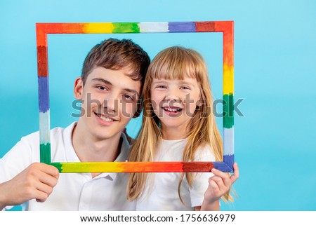 a 5-year-old girl and a teenage boy look at the camera through a colored frame and smile, on a blue background, copy space