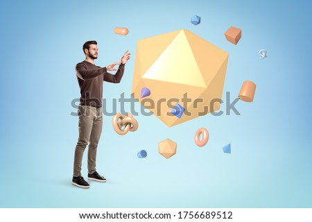 Young man in standing and trying to manipulate a huge yellow icosahedron floating in air with lots of other small geometric objects on blue background. Spatial intelligence. Education and learning. Royalty-Free Stock Photo #1756689512