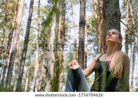a girl in a forest sits on a tree, with headphones listening to music. rest at nature. in the background blurred silhouettes of trees. digital detox. Mental health. Positive emotion. Royalty-Free Stock Photo #1756704626