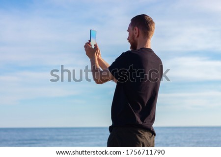 Bearded man in black t-shirt on a sea and clear blue sky landscape background with smartphone in hands taking a picture. Young guy photographing nature on cell phone, active lifestyle travel concept.