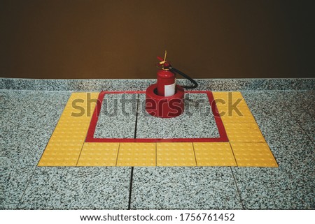 A red powder fire extinguisher indoors of an airport or a mall in a special yellow zone made of tactile tiles on the marble floor near a brown wall
