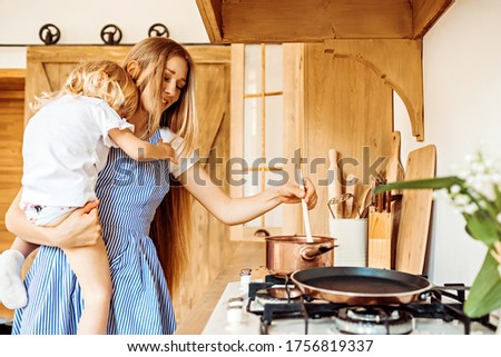 Young mother or babysitter cooks food with a little girl in her arms in the kitchen at home.