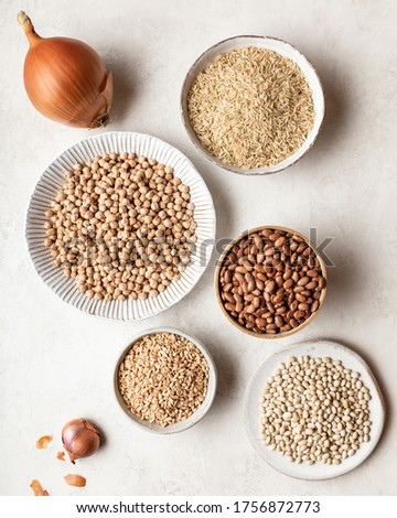 Multiple bowls are filled with a variety of pantry staples. Rice, barley, and several types of beans sit in individual bowls on a neutral background, along with an onion and shallot. Royalty-Free Stock Photo #1756872773