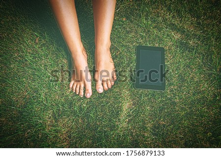 Tablet PC is lying on grass near the bare feet of female student. Shot from above. Faded look toned in vintage colours.