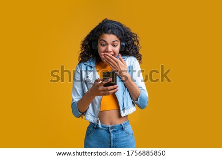 Excted dark skinned girl in wireless headset reading message on her mobile phone covering her mouth in amazement. Royalty-Free Stock Photo #1756885850