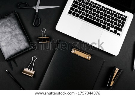 Top view of a workspace with laptop computer keyboard, notebook, pen, clipboard and clips on black background. Close up, copy space, background, flat lay
