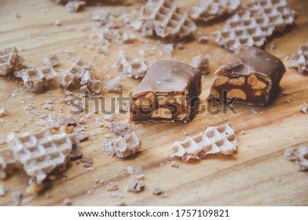 Close up picture of chocolate candy cut on parts on wooden background with waffles on sides, bar with peanuts and nougat covered with milk chocolate, as home made food photography and concept
