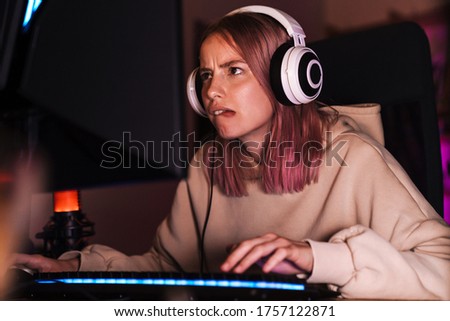 Image of confused focused girl in headphones playing video game on computer while sitting at table indoors
