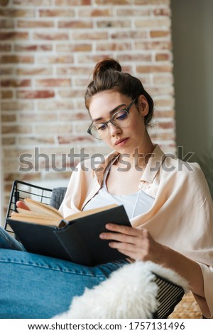 Image of focused beautiful woman in eyeglasses reading book while sitting in armchair at home Royalty-Free Stock Photo #1757131496