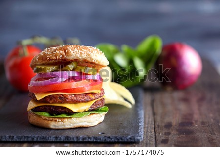 Vegetarian cheeseburger made with two meat substitute patties, slices of melted cheese, onions, pickles, lettuce, & tomato on a fresh sesame seed bun over a rustic dark background.  Blurred Background