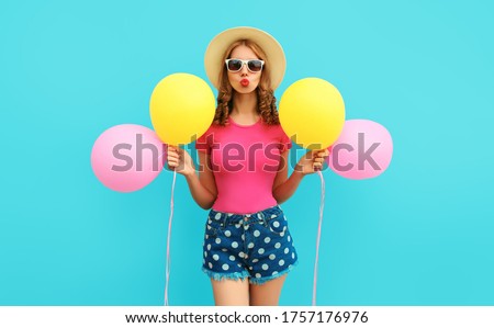 Summer colorful cheerful image of cute young woman with yellow pink balloons having fun wearing a shorts and straw hat on blue wall background