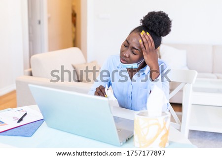 Attractive black young businesswoman sitting alone in her office and coughing as she suffers from a cold. Medical mask and hand disinfectant and stressed woman.