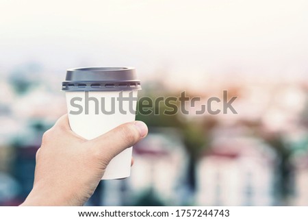hand holding paper cup of take away drinking coffee on city background natural morning sunlight. space Place for your text or logo.