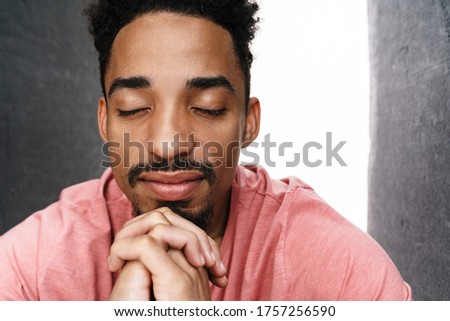 Photo of calm african american man with eyes closed posing on camera isolated over grey wall with white light