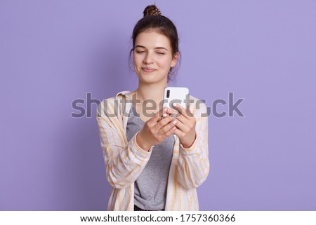Young happy smiling female wearing casual attire, posing against lilac wall with smart phone in hands, taking selfie picture, looking smiling at device screen.