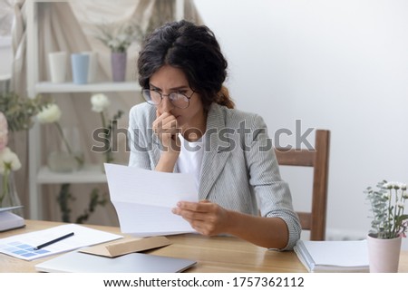 Pensive young Caucasian woman in glasses read message in paper correspondence, think analyze, thoughtful female sit at desk feel stressed confused consider unpleasant notice in paperwork letter Royalty-Free Stock Photo #1757362112