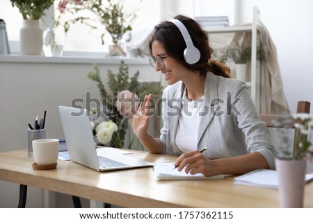 Smiling young Caucasian woman in headphones take online educational course or training on laptop from home, happy female in wireless headset wave to camera, talk on webcam video call on computer Royalty-Free Stock Photo #1757362115