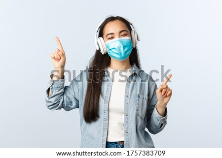 Social distancing lifestyle, covid-19 pandemic and self-isolation leisure concept. Happy upbeat asian girl in medical mask, listening music in wireless headphones and dancing