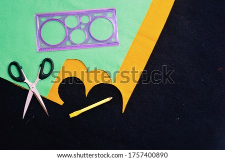 Top view of colorful felt textile material cut out for craft project with accessories like scissor, ruler and fabric marker on black felt background. 