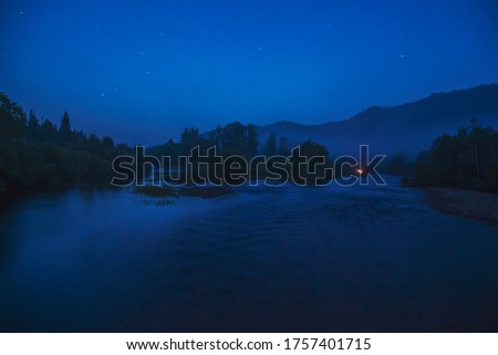 Night landscape: a small flat river, a dense coniferous forest and a tourist tent with bright light. Photos from a trip to Russia in the summer.