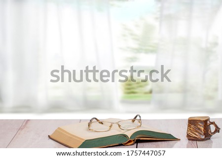 Books backgrounds. Closeup of an antique book with reading glasses on it and a brown clay mug on a pink desk in front of abstract blurred light green white curtain background. Teachers day.