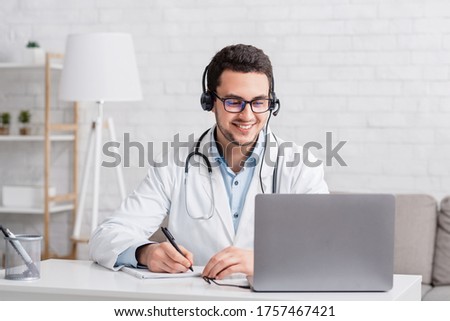 Smiling doctor in white coat with stethoscope and headphones, takes notes in notebook looks in laptop on online lesson, free space