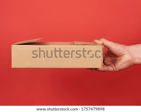 male hand holds a brown cardboard box on a red background, concept of goods delivery