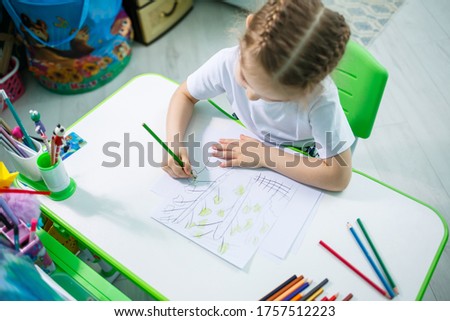 Portrait of a cute little girl looking at the camera and smiling while drawing pictures or doing homework, sitting at a table in the home interior, copy space