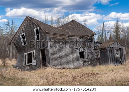 An abandoned house that is collapsing in a field. Windows and door broken out, no trespassing sign on front. Blue sky with clouds.