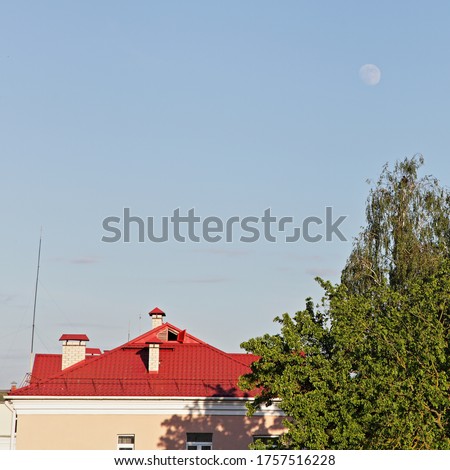 A full moon in a clear blue sky above the old red tile roof of a house and the green top of a tree on a summer evening, picture with copy space for text