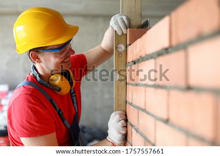 Close-up of bricklayer using spirit level to check new red brick wall. Professional worker in yellow helmet smiling at work. Construction site and renovation concept Royalty-Free Stock Photo #1757557661