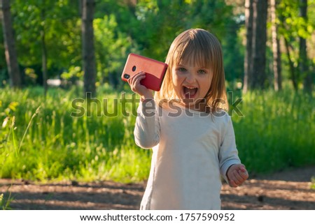 A little girl of 2 years old is holding a red smartphone in her hand with a triumphant, joyful expression on her face. Talking on the phone, emotions, selfies. Children and gadgets.