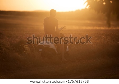 A young man sits on a motobike near a lavender field at sunset.
Image with noise effects and selective focus.
