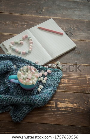 A cup of a common drink with marshmallows on a knitted scarf, a heart made of small sweets, an open notebook,  pink pencil. wooden dark background. Concept of love, make wishes. Copy space. Top view. 
