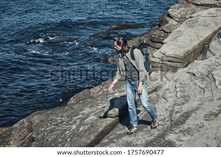 young long-haired man with a backpack and a photo tripod explores the rocky sea coast. Travel and outdoor concept.