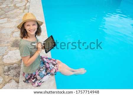 Happy joyful woman in summer outfit sitting at poolside, using tablet with pen, swinging legs in blue water, looking at camera. Outdoor shot, day. Work on vacation or technology concept
