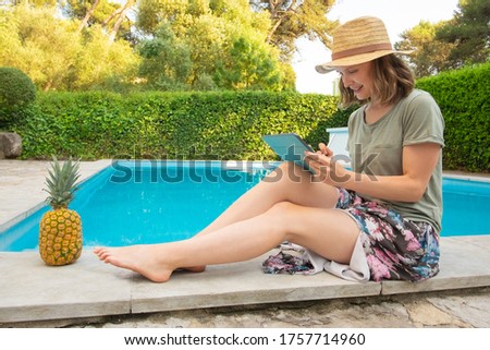 Happy focused woman sitting at swimming pool, using tablet and stylus, drawing pineapple. Outdoor shot, side view. Summer or working outdoors concept