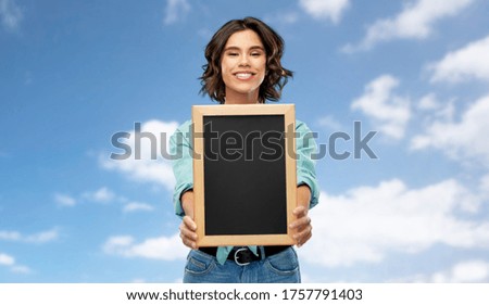people concept - portrait of happy smiling young woman in turquoise shirt showing black chalkboard over blue sky and clouds background